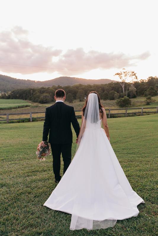 KWH real bride Hannah and Reece walking in a field. She wears the ethereal Blake Camille gown, a modern strapless aline wedding dress.