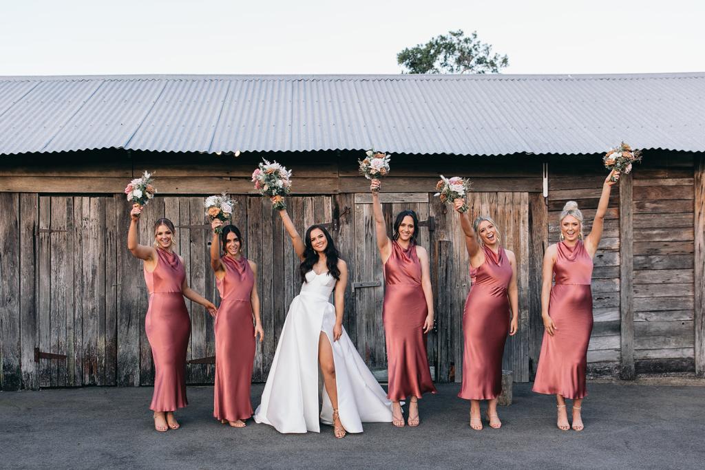 KWH real bride Hannah and her bridesmaids lifting their bouquets in the air. She wears the Blake Camille gown, a modern strapless aline wedding dress.