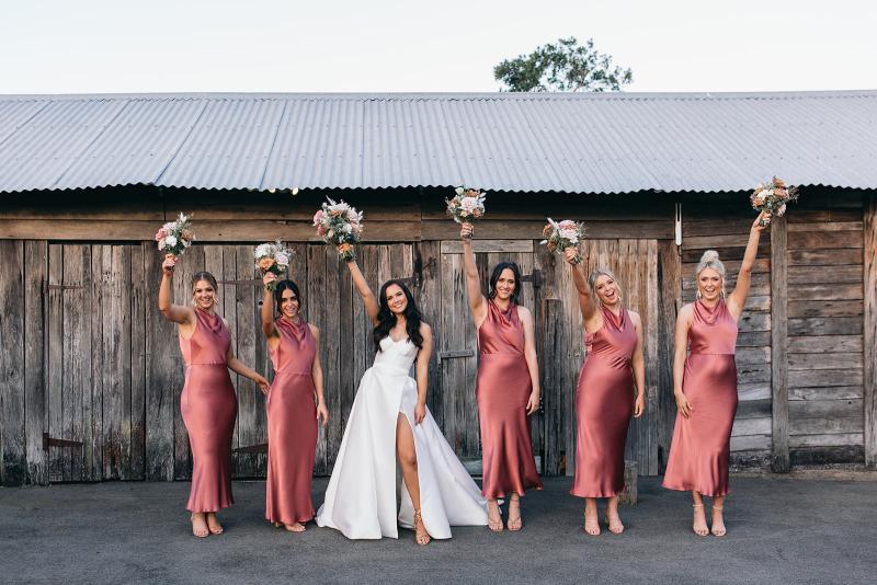 KWH real bride Hannah and her bridesmaids lifting their bouquets in the air. She wears the Blake Camille gown, a modern strapless aline wedding dress.