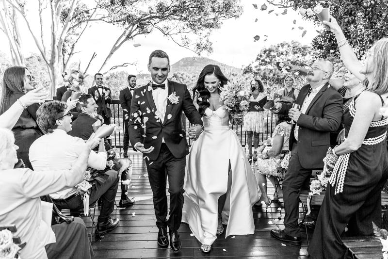 B&W image of KWH real bride Hannah and Reece walking down the aisle as she wears the Blake Camille gown, a modern strapless aline wedding dress.