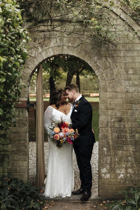 KWH real bride Nalini and Michael hug underneath a stone arch. She wears the Celine gown, v-neck fit and flare beaded wedding dress.