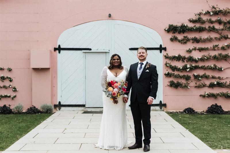 KWH real bride Nalini and Michael stand in front of a baby pink and blue Mona farm. She wears the Celine gown, v-neck fit and flare beaded wedding dress.