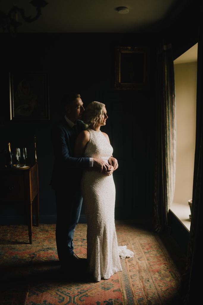 KWH real bride Laura and David stand by the glow of the window. She wears the Cindy gown, a halter neck sequin wedding dress.