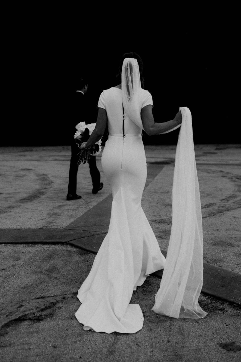 B&W image of KWH real bride Jacqui walking by a black wall. She wears the Clarissa gown, a high back fit and flare cap sleeve wedding dress with the long peal veil.