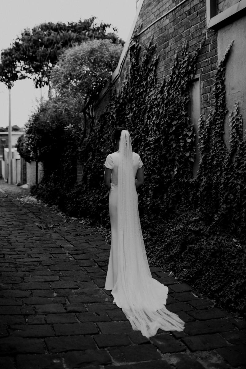 B&W image of KWH real bride Jacqui standing by an ivy wall. She wears the Clarissa gown, a high back fit and flare cap sleeve wedding dress.
