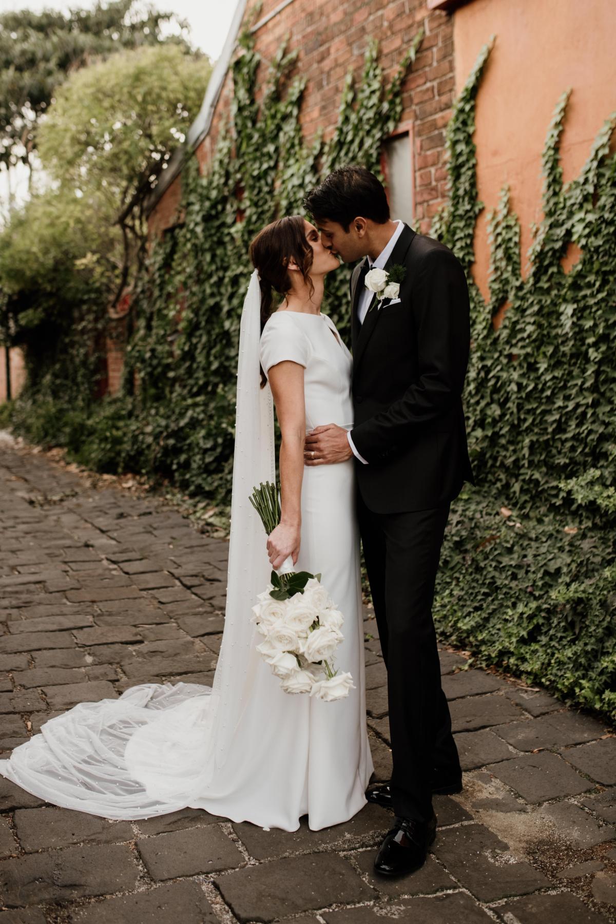 KWH real bride Jacqui and Chris kiss by the ivy wall. She wears the long pearl veil with her Clarissa gown, a fit and flare wedding dress with high neck cap sleeve.