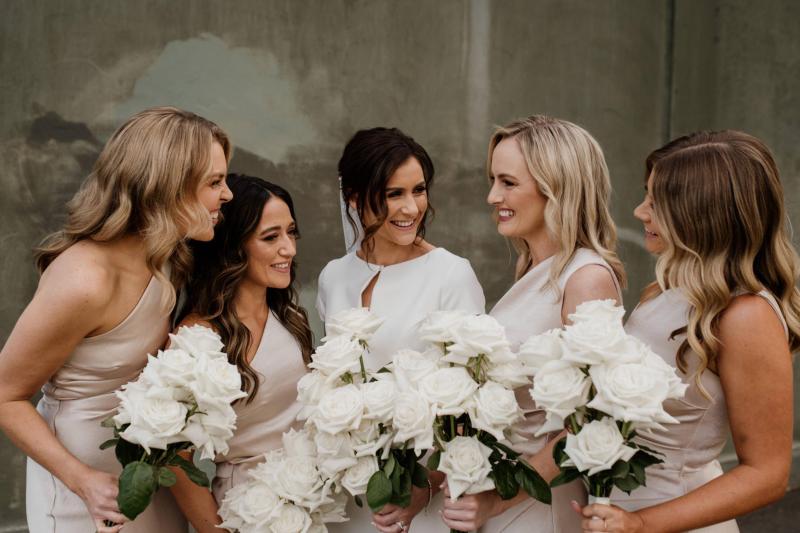 B&W image of KWH real bride Jacqui walking by a black wall. She wears the Clarissa gown, a high back fit and flare cap sleeve wedding dress with the long peal veil.