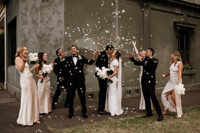 B&W image of KWH real bride Jacqui walking by a black wall. She wears the Clarissa gown, a high back fit and flare cap sleeve wedding dress with the long peal veil.