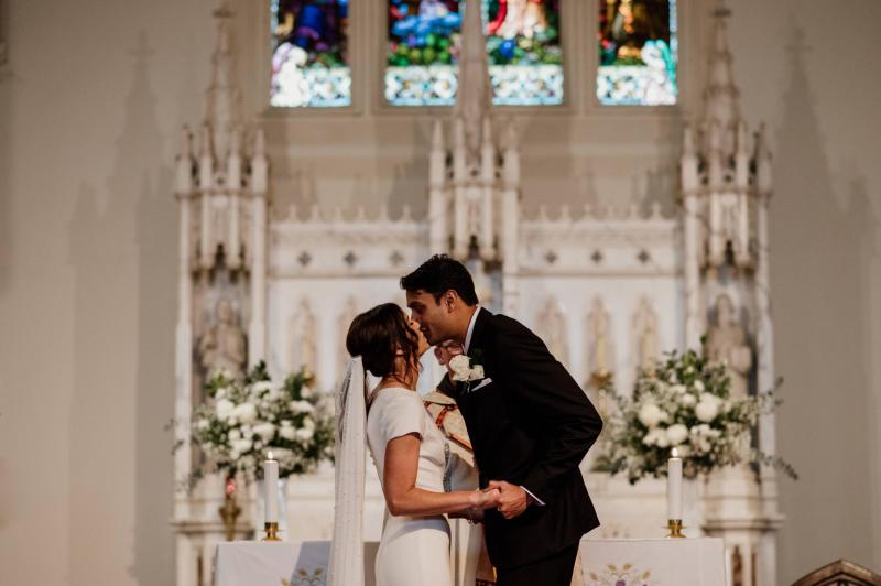 KHW real bride Jacqui and Chris kiss at the church altar. She wears the Clarissa gown, a high back fit and flare cap sleeve wedding dress.