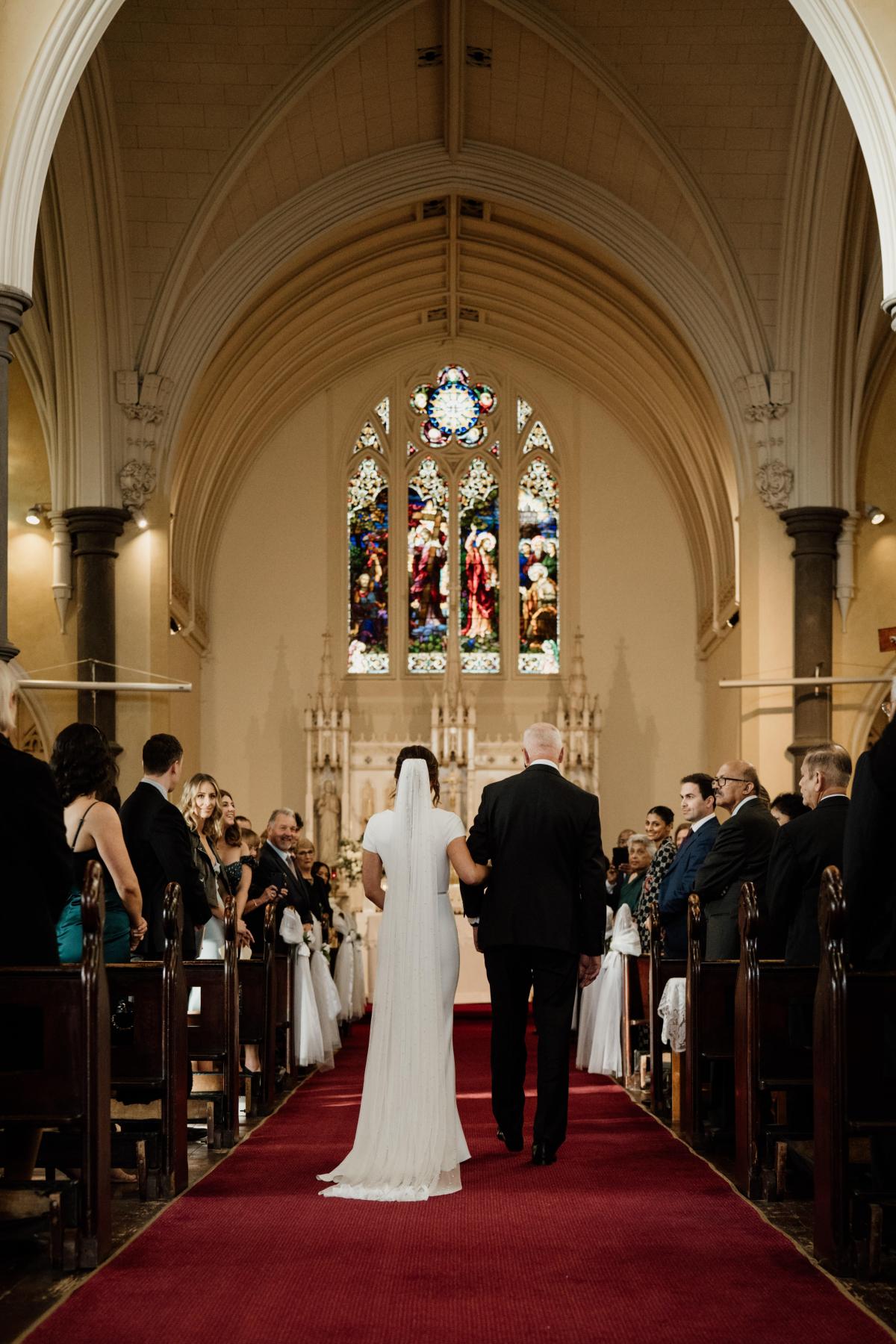 KWH real bride Jacqui is walked down the aisle by her dad at her church wedding. She wears the Clarissa gown, a high back fit and flare cap sleeve wedding dress.