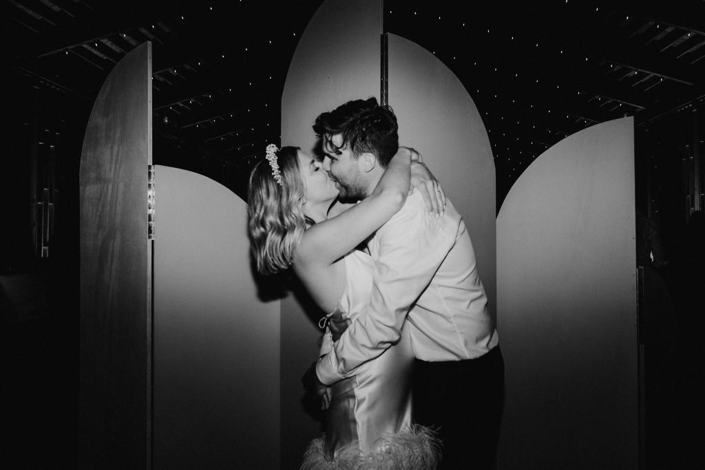 B&W image of KWH real bride Kasey kissing Brian at their recption. She wears the Sherry mini, a simple short wedding dress with open back and feather detail.