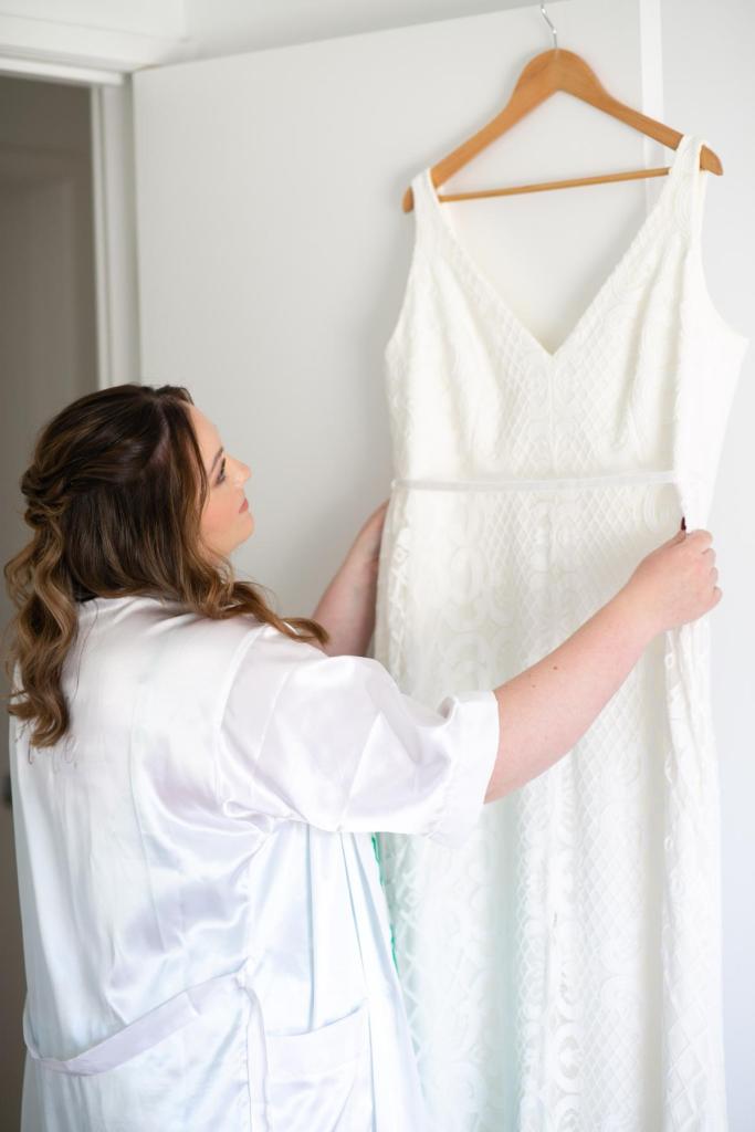 KWH real bride Meg looks at her Bobby gown hanging up. Bobby is a fit and flare gown made from lace.
