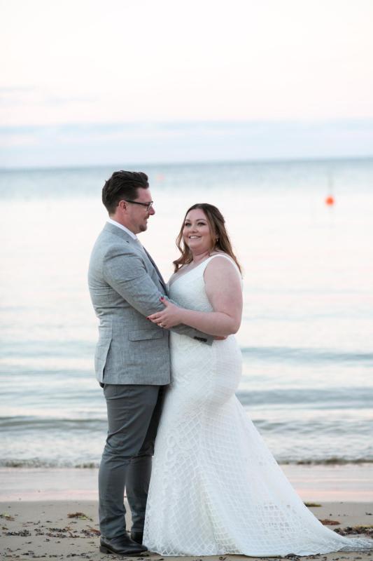 KWH real bride Meg and tom hug on the beach. She wears the bobby gown, a modern lace fit and flare wedding dress.