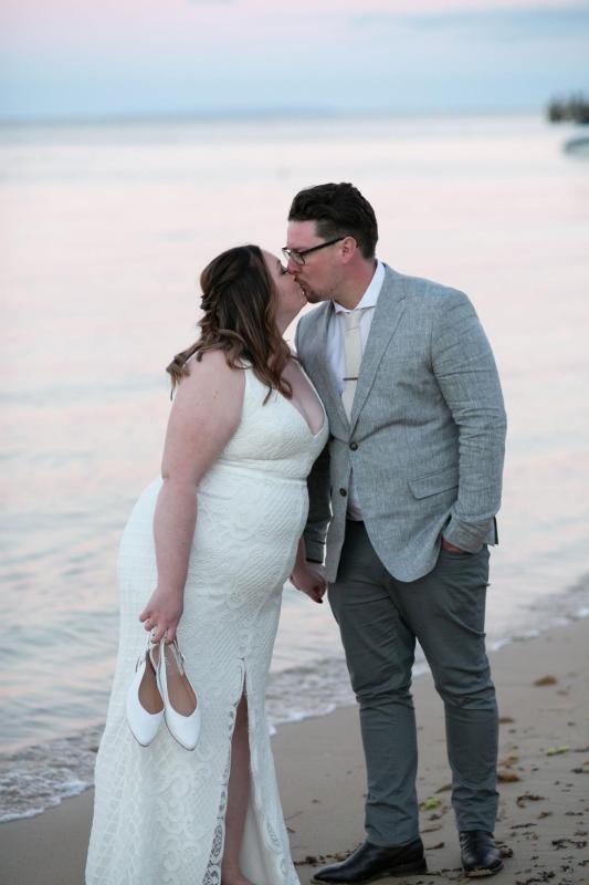 KWH real bride Meg kisses Tom on the beach. She wears the effortless Bobby gown, a modern fit and flare wedding dress.