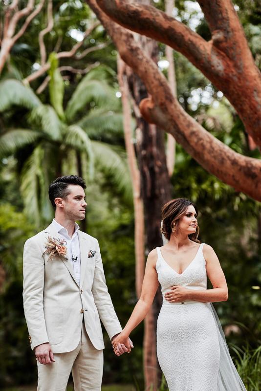 KWH real bride Emma and Will hold hands in the lush greenery as she stands in her Rylie gown, a modern lace wedding dress with vneck.
