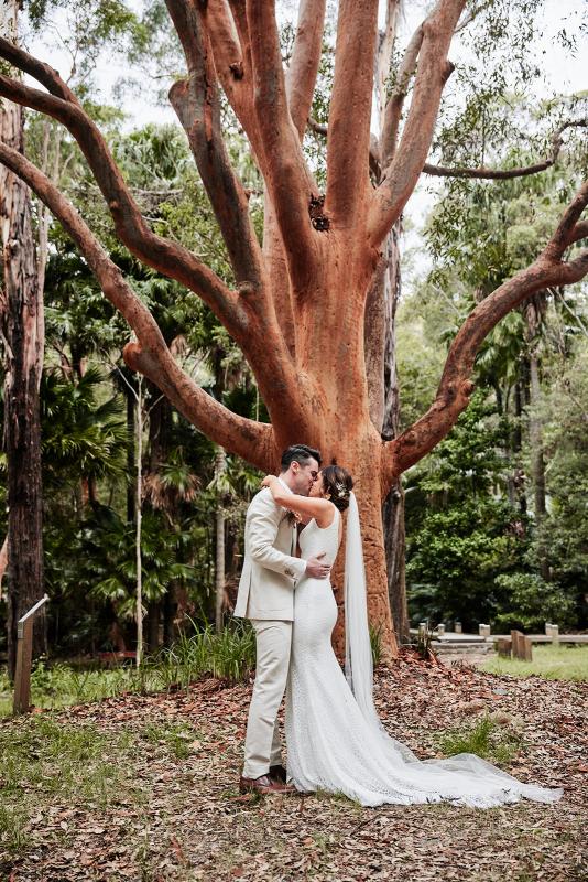 KWH real bride Emma and Will kiss in front of a huge tree. She wears the Rylie gown, a modern lace fit and flare wedding dress.