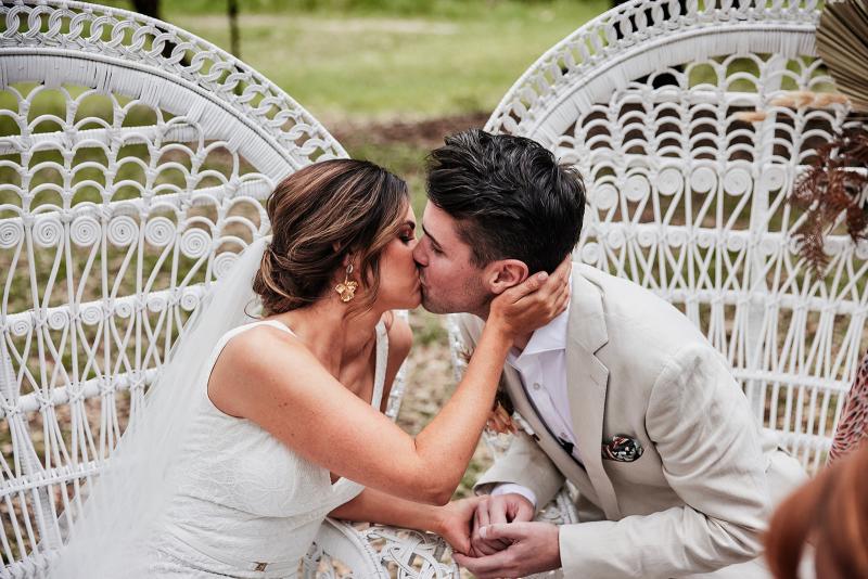 KWH real bride Emma and Will kiss in their bridal chairs. She wears the Rylie gown, a sophisticated lace fit and flare wedding dress.