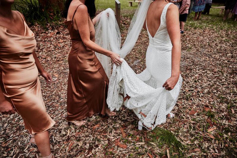 KWH real bride Emma walks as her bridesmaids hold the train of her Rylie dress up. Rylie is a modern lace fit and flare wedding dress.