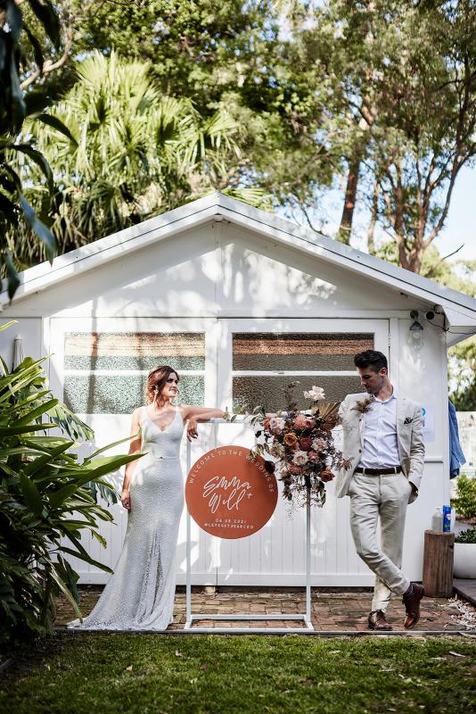 KWH real bride Emma and Will stand by their wedidng signage. She wears the Rylie gown, a modern lace fit and flare wedding dress.