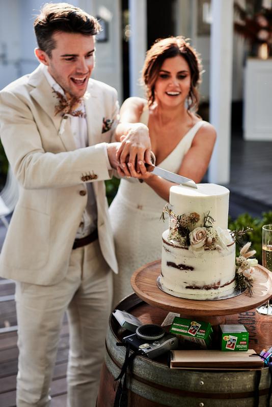KWH real bride Emma and Will cut the cake. She wears the comfortable Rylie gown, a modern lace fit and flare wedding dress.