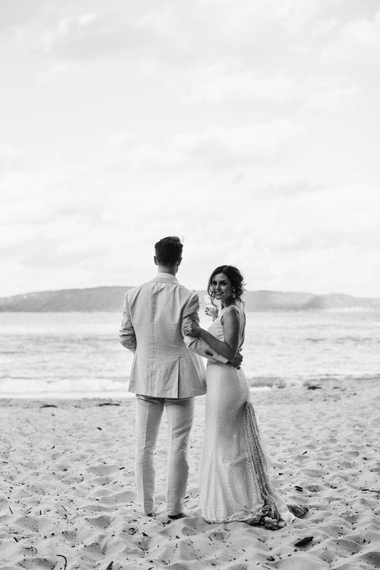 B&W image of KWH real bride Emma and Will standing on the beach. She wears the effortless Rylie gown, a modern lace fit and flare wedding dress with long train.
