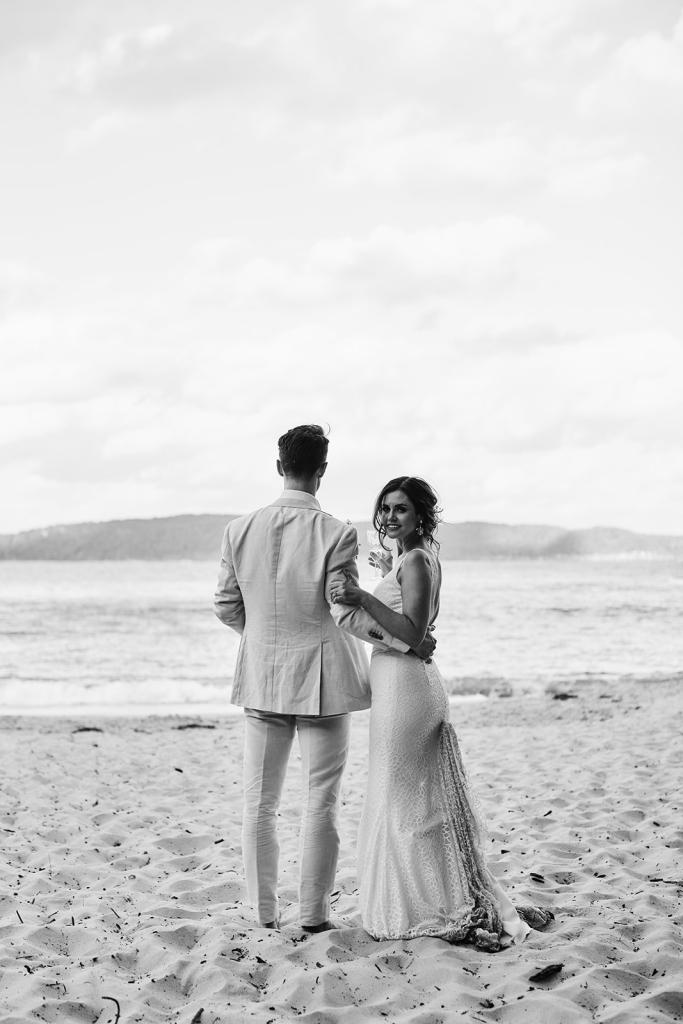 B&W image of KWH real bride Emma and Will standing on the beach. She wears the effortless Rylie gown, a modern lace fit and flare wedding dress with long train.