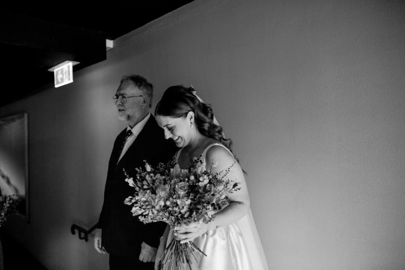 B&W image of KWH real bride Tara walking down the aisle with her father. She wears the grand Taryn Camille gown, a sophisticated aline wedding dress with U-shaped neckline.