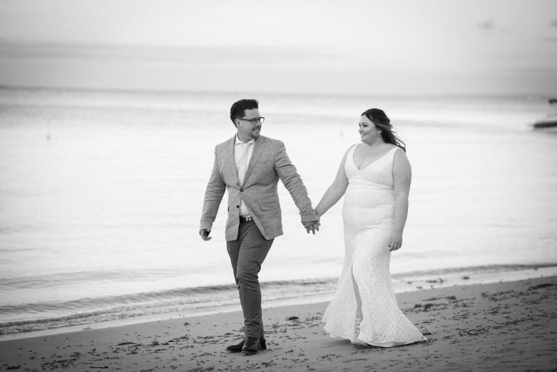 KWH real bride Meg and Tom walk down the beach together. She wears the Bobby gown, a modern fit and flare lace wedding dress.