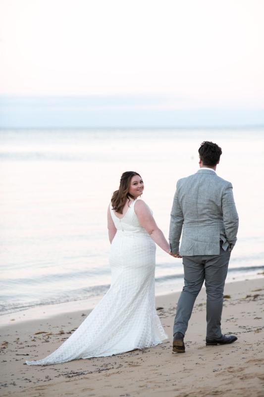 KWH real bride Meg walks down the beach with Tom in her Bobby gown, a modern fit and flare lace wedding dress.