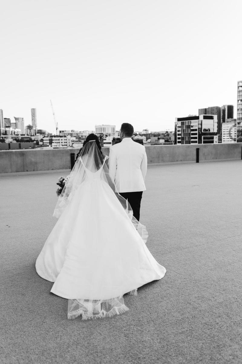 B&W image of KWH Real bride Josey and Brad walking toward the city skyline. She wears the Taryn Camille gown, a modern aline wedding dress with long train.