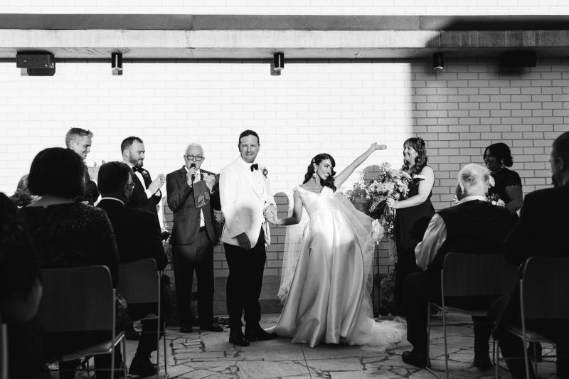 B&W image of KWH real bride Josey and Brad celebrating at their ceremony. She wears the Taryn Camille gown, a timeless aline wedding dress with U-shaped neckline.