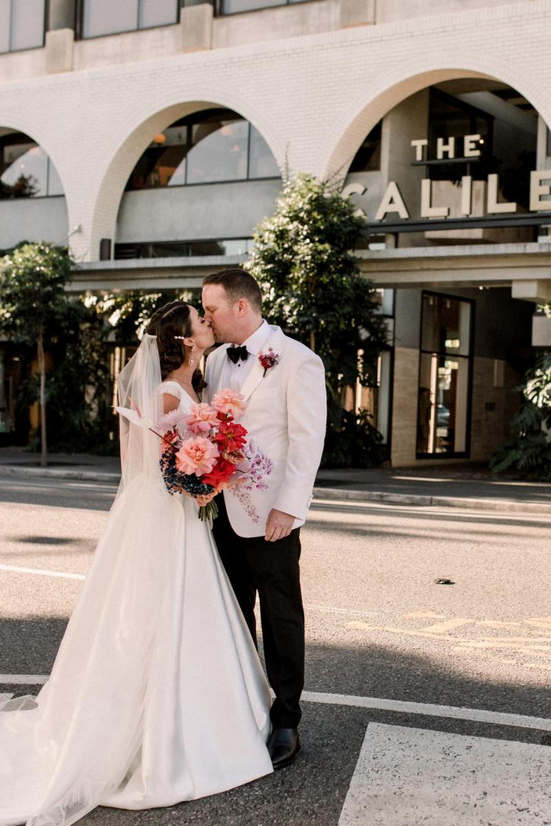 KWH real bride Josey and Brad kiss in front of the Calile Hotel. She wears the glam Taryn Camille gown, a modern aline wedding dress with U-shaped wedding dress.