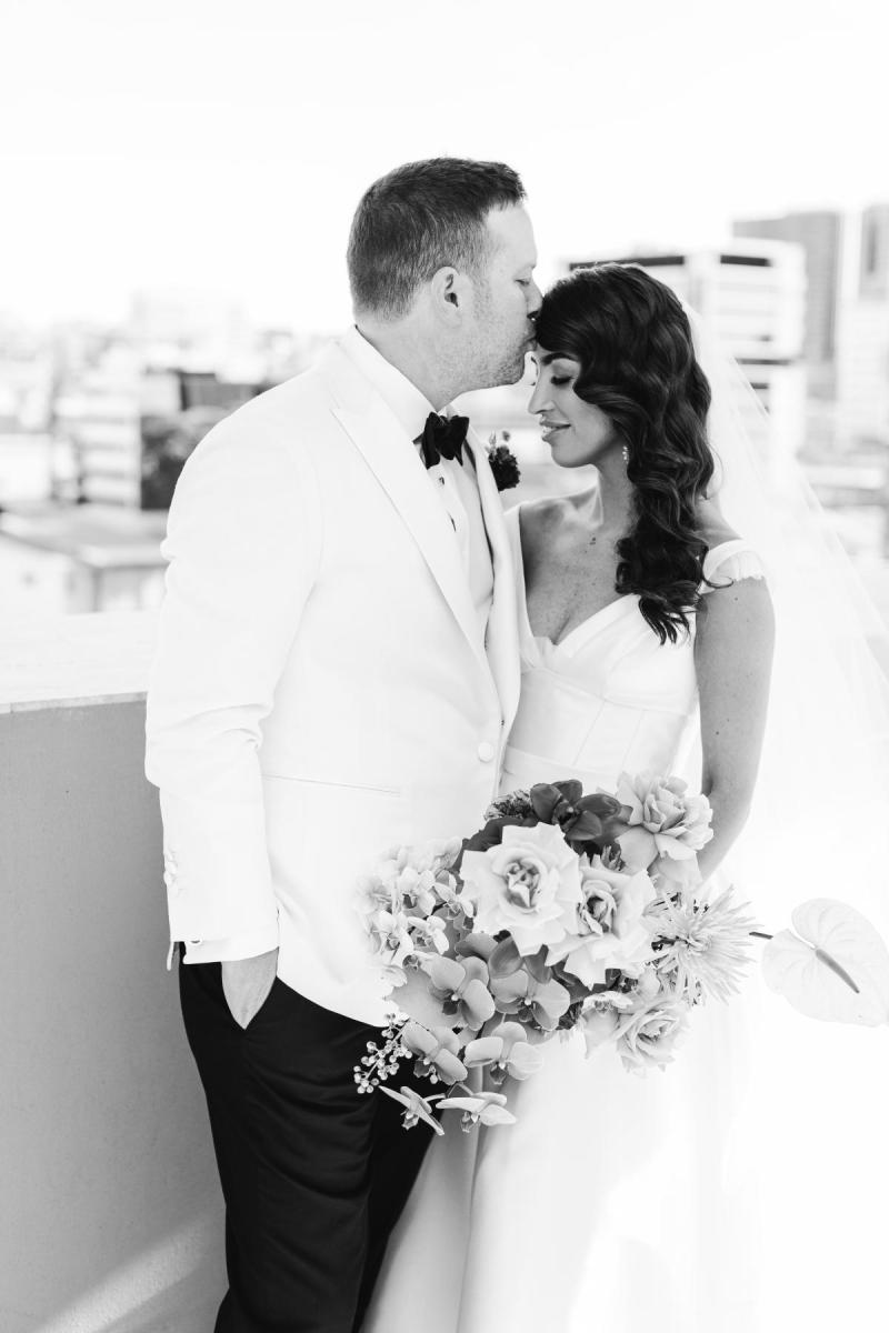 B&W image of KWH Real bride Josey getting a forehead kiss by Brad. She wears the glamorous Taryn Camille gown, a modern aline wedding dress with straps.