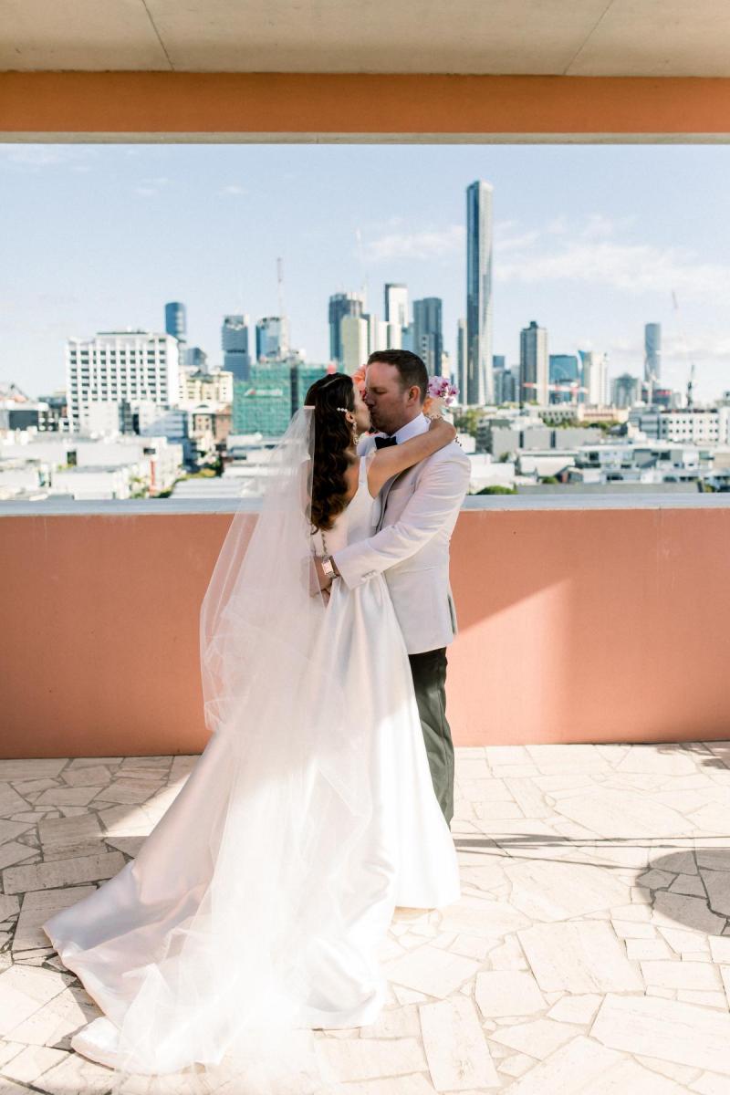 KWH real bride Josey and Brad kiss in front of the skyline as she wears the grand Taryn Camille gown, a modern aline wedding dress with U-shaped neckline.