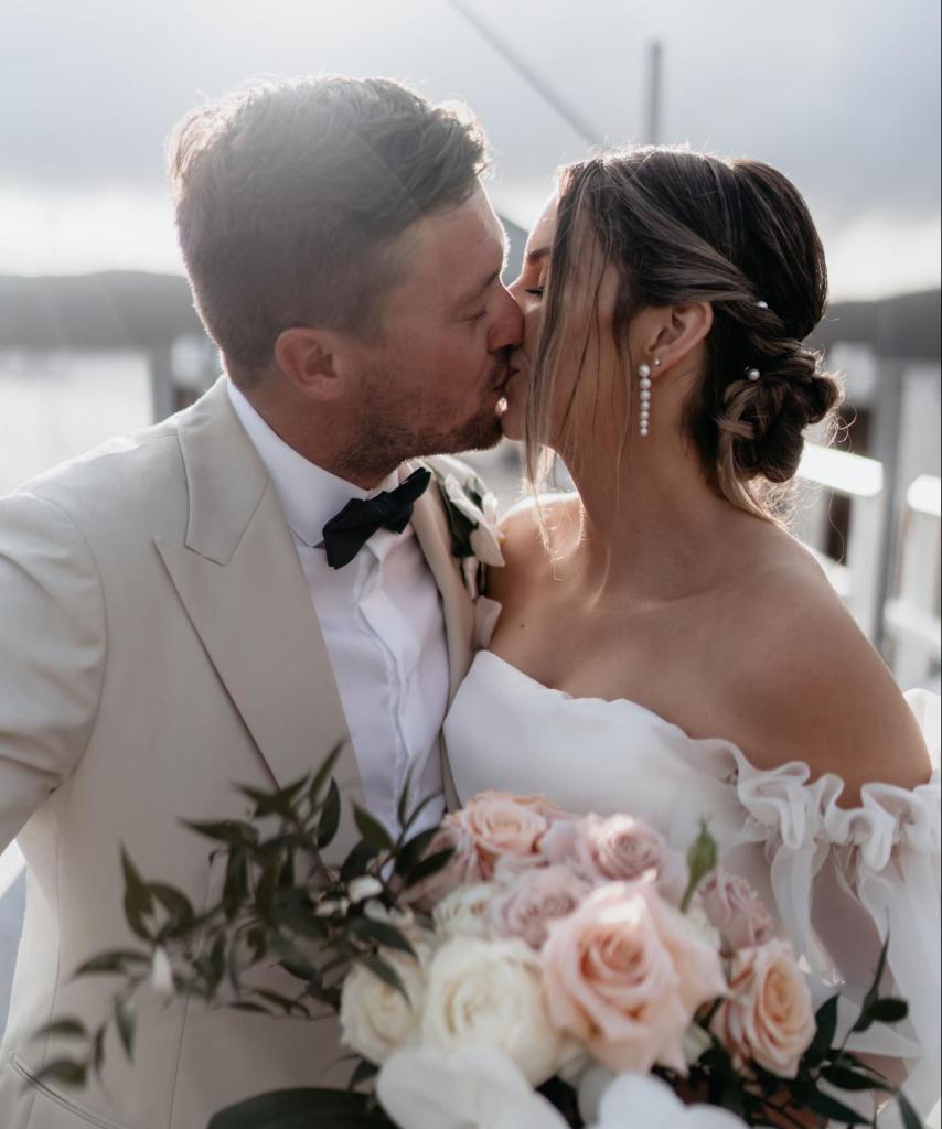 CATALINA - Karen Willis Holmes - Jessica & Nick - Bride & Groom Kiss, while bride holds her peachy tones flower bouquet, with a touch of off white elements