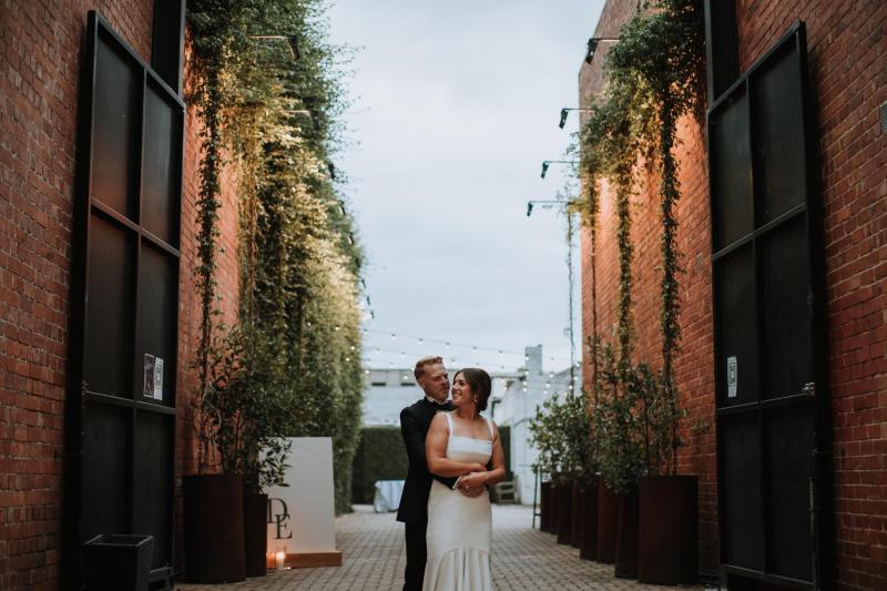 Newly married couple, Emily and Dan stand in the alley at Butler Lane. Real bride Emily wears the simple Violet gown by KWH.