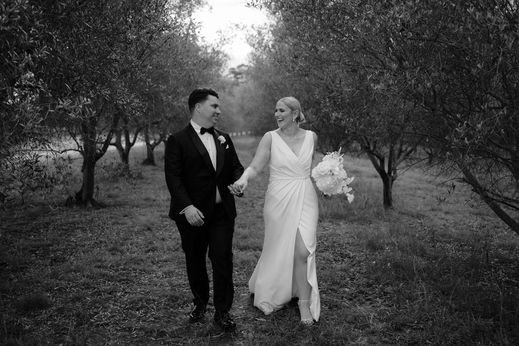 Nikki-Karen Willis Holmes- Bride and groom seen holding hands in a timeless vineyard full of views and bride holding off white flower bouquet