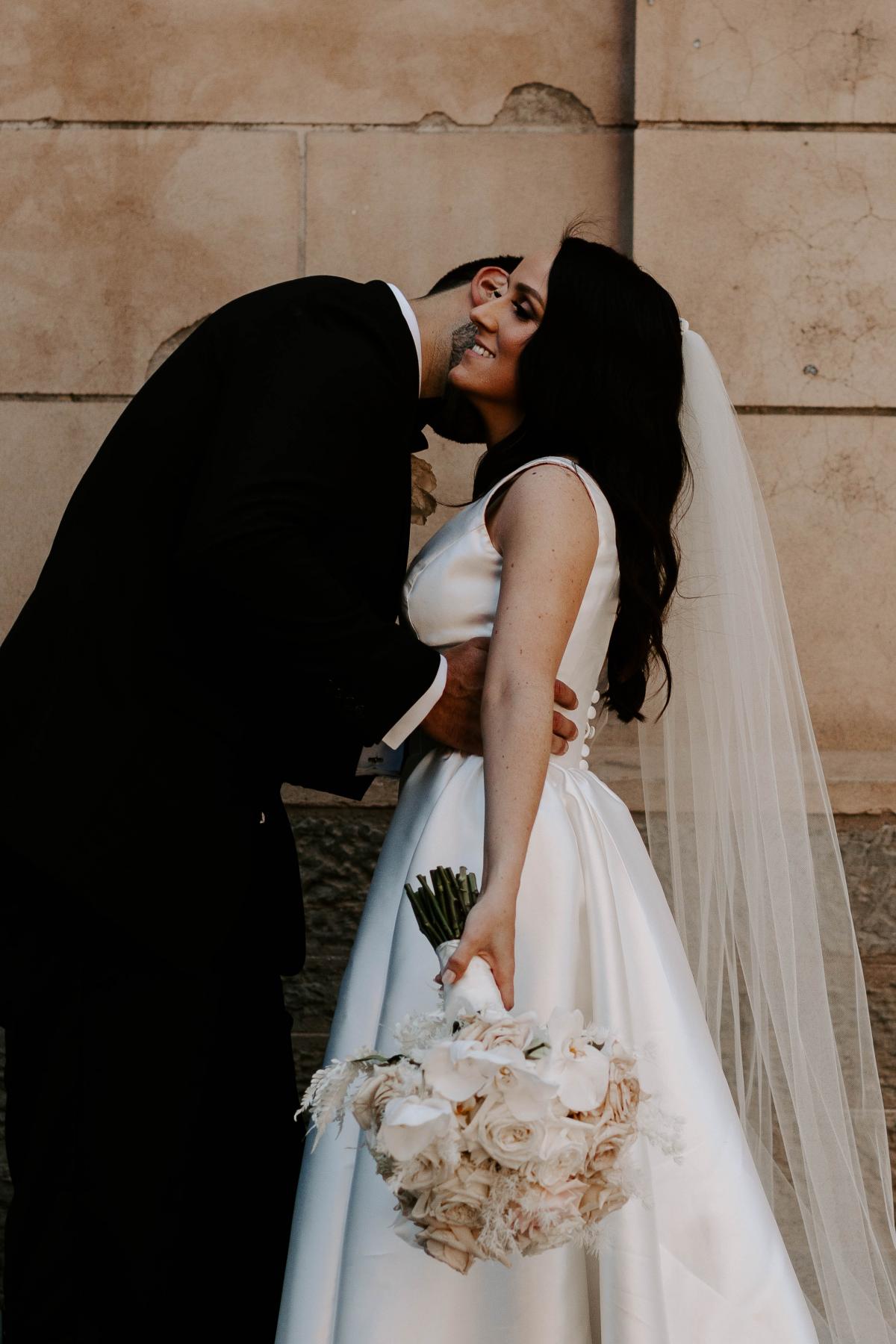 KWH real bride Shenea and Lain kissing by the sandstone wall. She wears the Taryn Camille gown with U-shaped bust, tight waist, and a-line skirt with pockets.