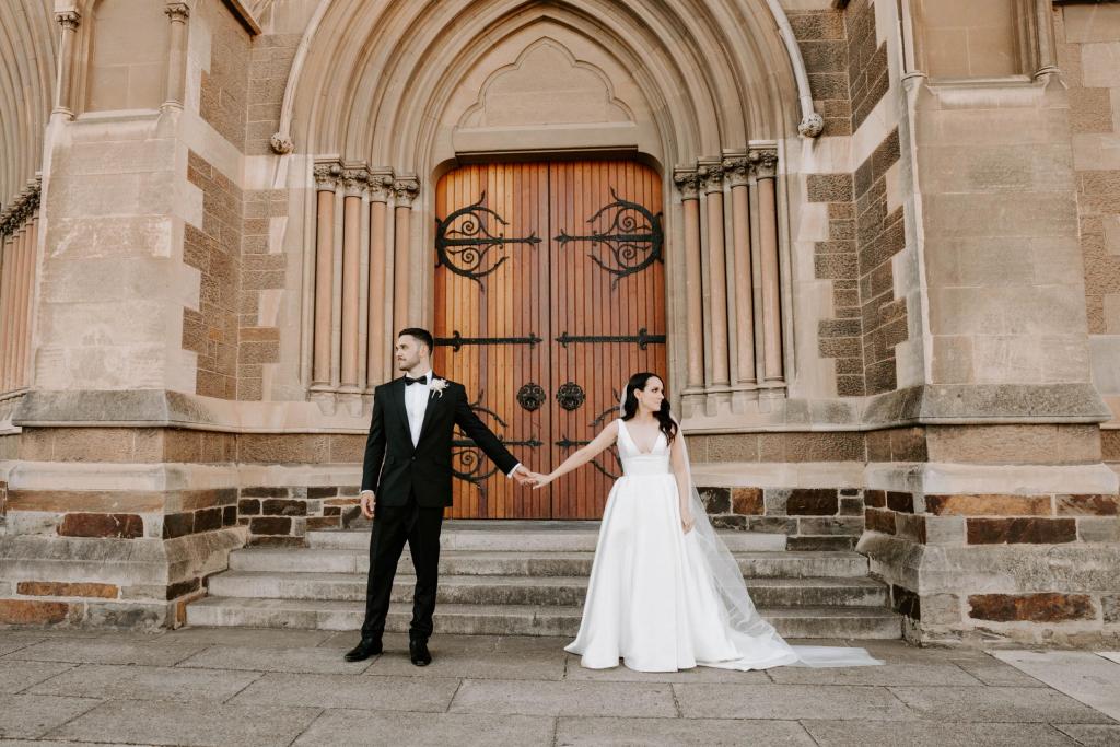 Real bride Shenea and Lain hold hands in front of the chapel doors as she dons the modern Taryn Camille gown from KWH, which features a U-shaped neckline and split in the skirt. . 