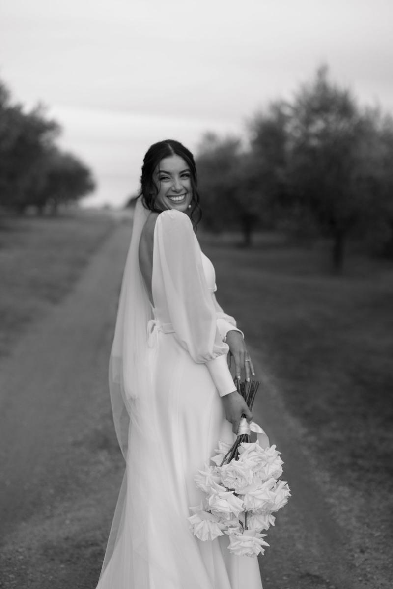 Brie Gown-Karen Willis Holmes - The bride Steph photographed smiling with bouquet, wearing the beautiful Brie gown from our Wild Hearts collection. Long sheer sleeves with button detail and low deep V back.