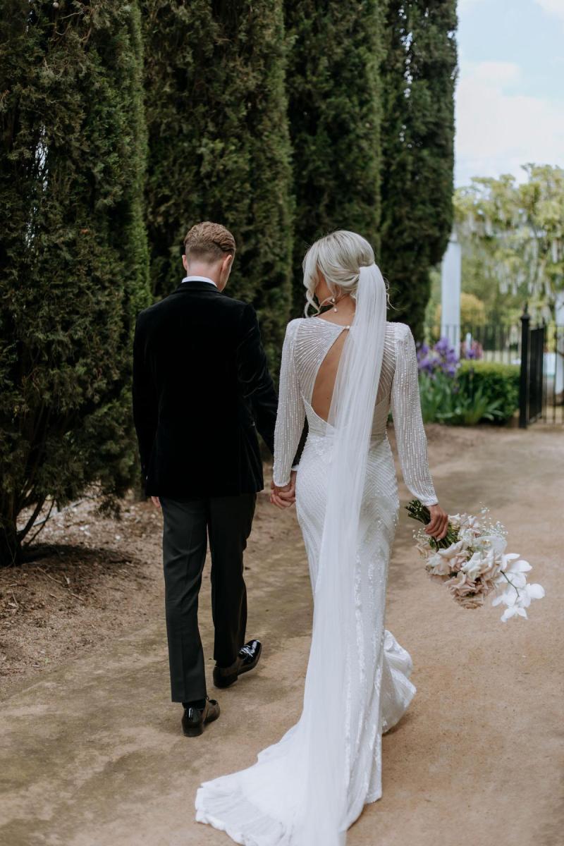 Perry - Karen Willis Homes - Laura & Ben - Bride and Groom seen holding hands while they walk through their wedding venue, surrounded by aces of beautfiul gardens and sophisticated structures.