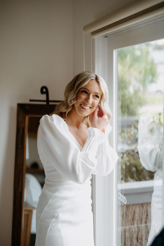 Brooklyn & Jade - Karen Willis Holmes - Stephanie & Ben - Bride Stephanie is seen getting ready before her special wedding day. Bride is looking out the window of her suite, while she wears the classic and sexy Brooklyn Jayde gown from the BESPOKE collection. Features a V-Neckline.