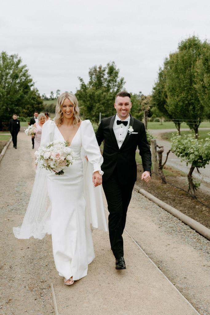 Brooklyn & Jade - Karen Willis Holmes - Stephanie & Ben - Bride and groom are seen holding hands while they adventure around their classic garden wedding venue in the Yarra valley. Bride is holding a soft pink and off white floral arrangement. Bride wears the Edgy and unique Brooklyn Jayde gown from the BESPOKE collection.