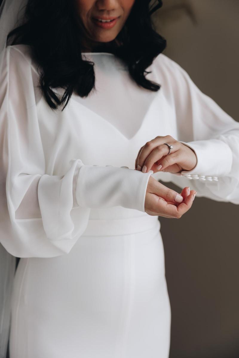 The Brie wedding dress by Karen Willis Holmes Bridal is a statement long sleeve sheer high neck gown with a low back, fitted skirt with a split. Brie features a v neck spaghetti strap bodice under the dreamy cuffed sleeves.