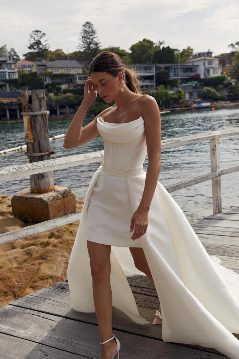 Styled with our Lizzie Train, the Evie & Neve Mini gown is classic with a modern flair. The Evie Bodice features a stunning scoop neckline and detachable drape to complement your décolletage with statement pipe boning lines for that touch of drama. The Neve Mini Skirt is the perfect match for brides who love a modern twist.
