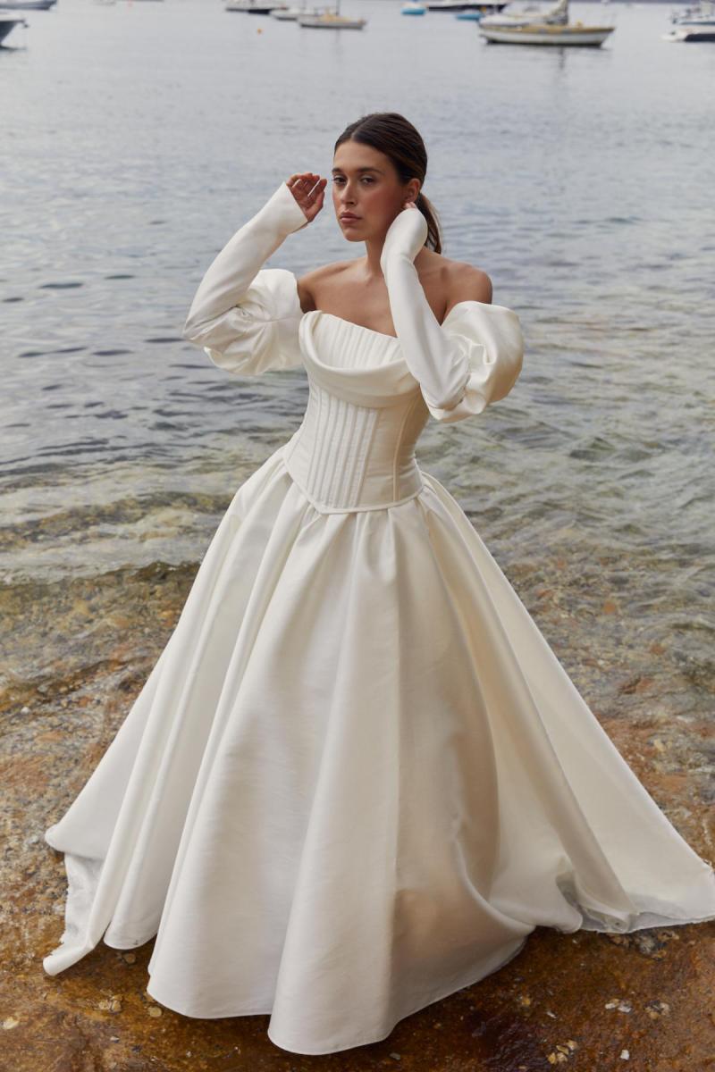 Roberta & Julianne is a classic wedding gown. The Roberta bodice features a long drop waistline with piping detail and detachable Evie drape and long puff Edwardian sleeves. Paired with the Julianne skirt which features a full ballgown silhouette.